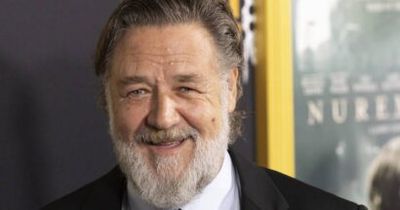 Smiling older man with a gray beard in a dark suit at a public event, with a backdrop behind him.
