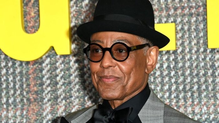 Older man in a gray suit with a black bow tie and round black glasses, wearing a black fedora at a red carpet backdrop.