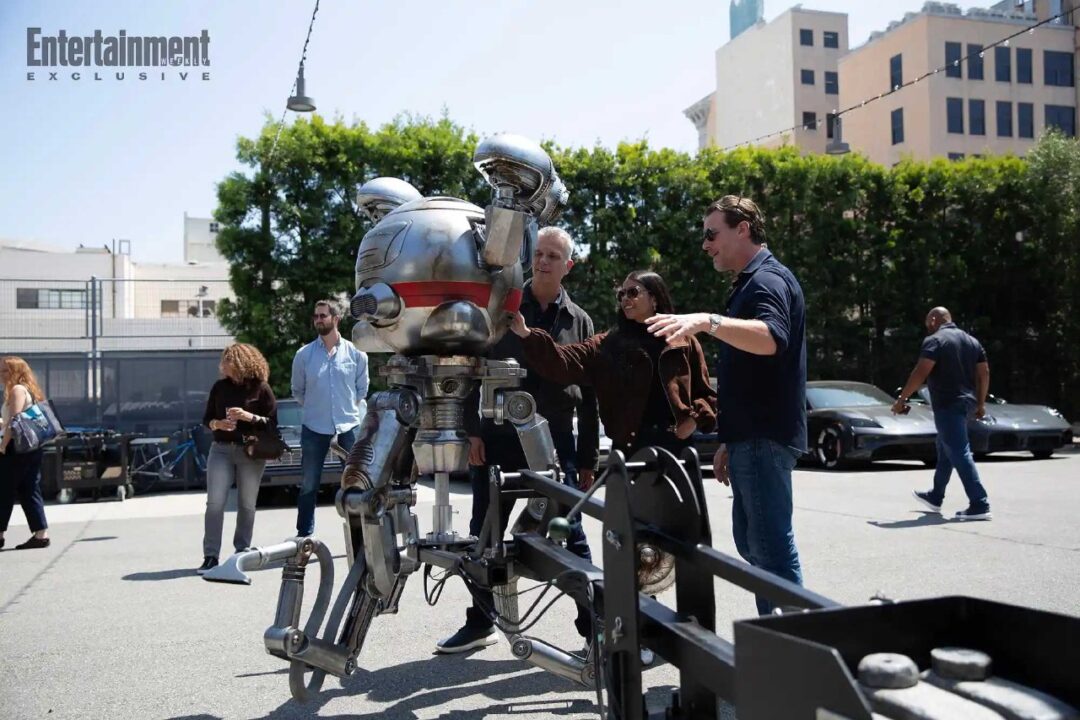 Group of People Examining a Large Silver Robot Prop on a Wheeled Frame Outdoors Entertainment Weekly Exclusive Logo Visible in Corner