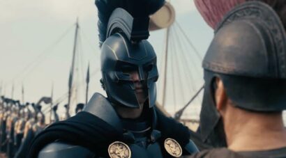 A armored warrior in a metallic helmet speaks to a fellow soldier on a historic battlefield with lined troops in the background