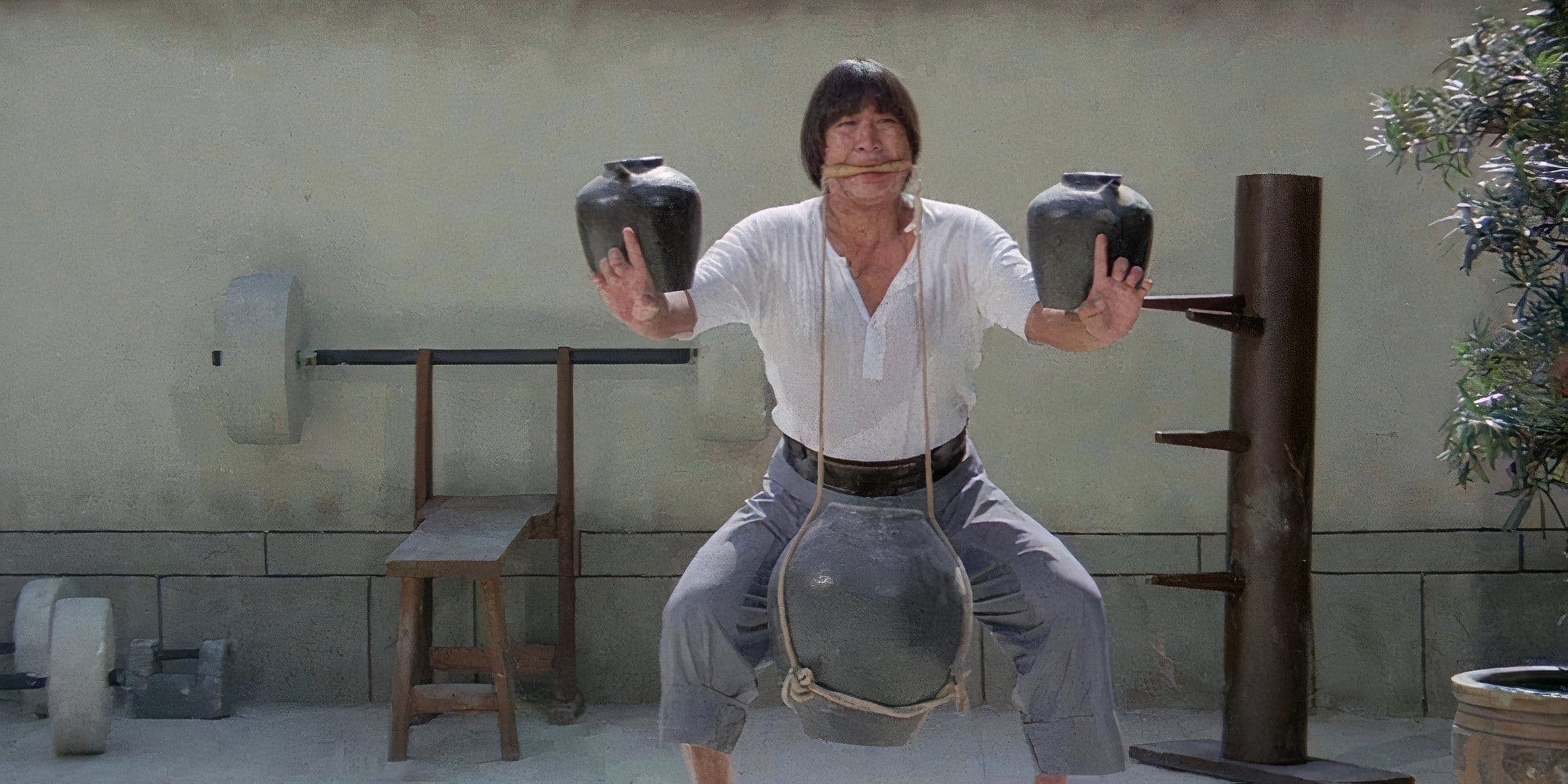 Man in a Light Shirt and Shorts Performing a Dramatic Strength Pose Holding Two Large Ceramic Jars in Each Hand Outdoors with a Heavy Weight Between His Legs