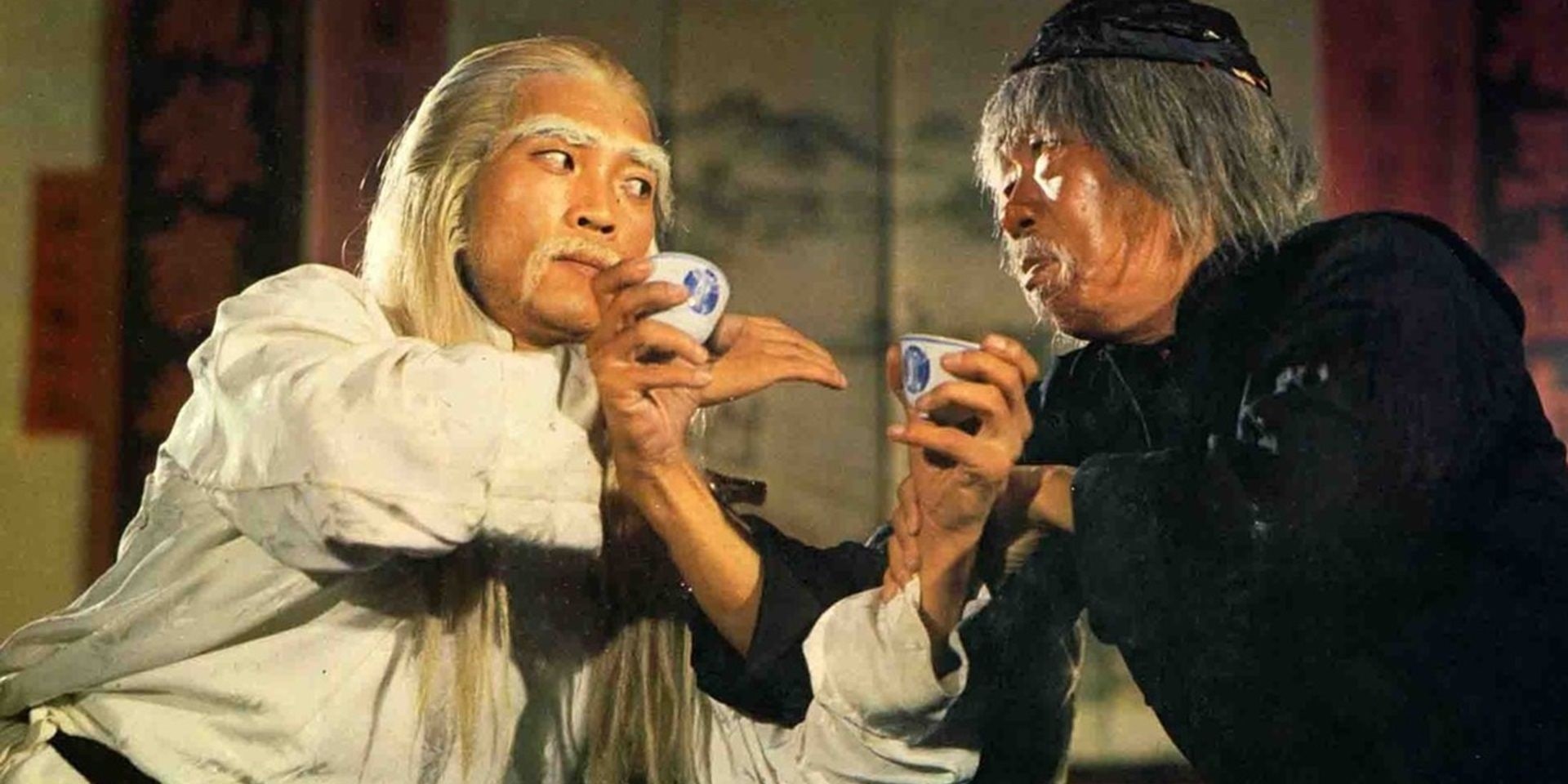 Two Costumed Older Men with White Hair Share Small Cups in a Tense Standoff Facing Each Other