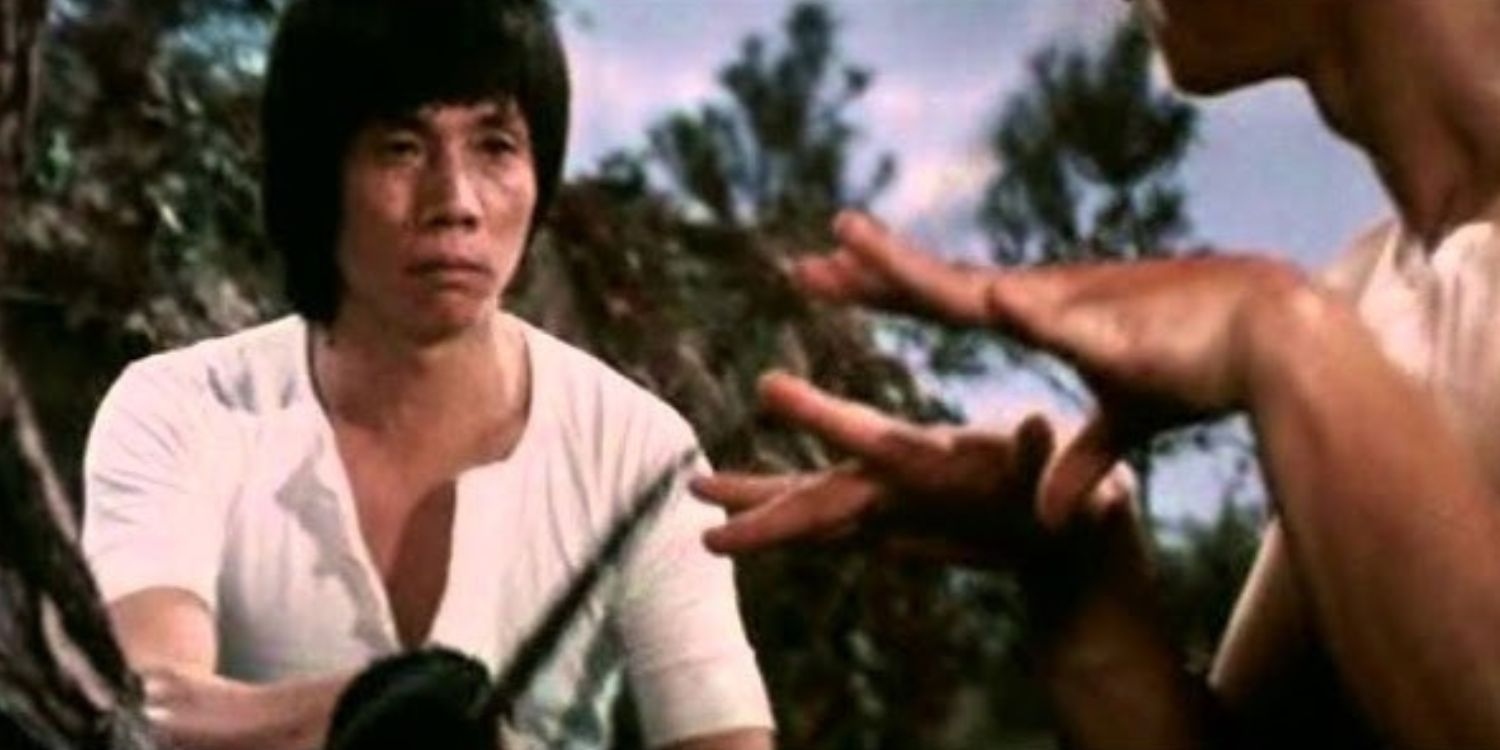 Man in a White Shirt Sits Outdoors with a Serious Expression As Another Person Extends a Hand Toward Him in a Tropical Setting