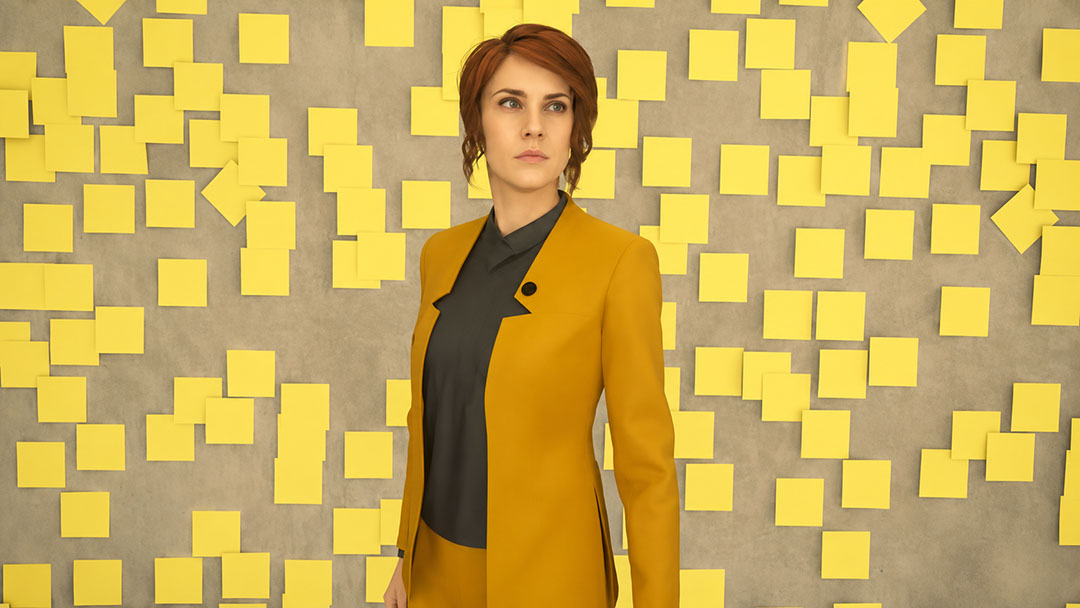 Control Woman with Short Reddish brown Hair in a Mustard Blazer over a Dark Blouse Standing in Front of a Gray Wall Covered with Yellow Sticky Notes