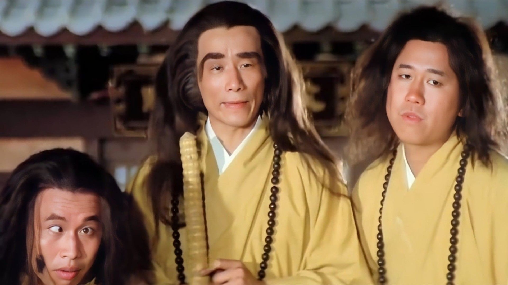 Three Men in Yellow Robes with Long Dark Hair and Bead Malas Stand Together in a Temple Setting Calm Expressions on Their Faces