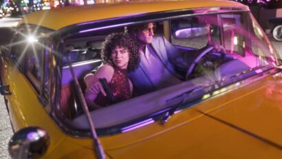 Yellow taxi at night with a man driving and a woman in the passenger seat holding a handgun, neon city lights reflected inside the car.