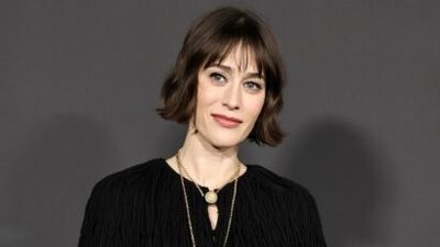 Woman with short brown hair wearing a black textured blouse and layered gold necklaces, against a gray background.