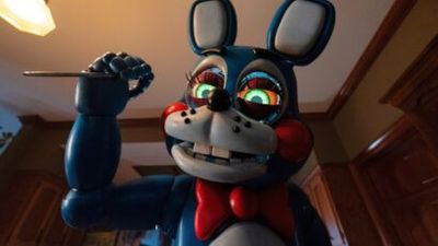 Blue animatronic bunny raises a knife, looking toward the viewer with glowing green eyes, red bowtie, indoors in a kitchen-like room.