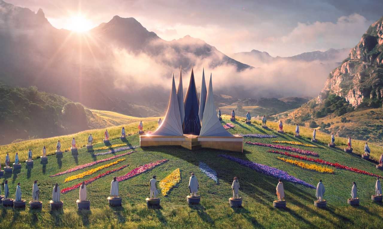 رده سنی جدیدترین فیلم Hunger Games مشخص شد | موبوگیم 1 Sunlit hillside sculpture garden with a central multi-spired white monument surrounded by a circle of statues on pedestals, and colorful flowerstrips across the slope with misty mountains in the distance.