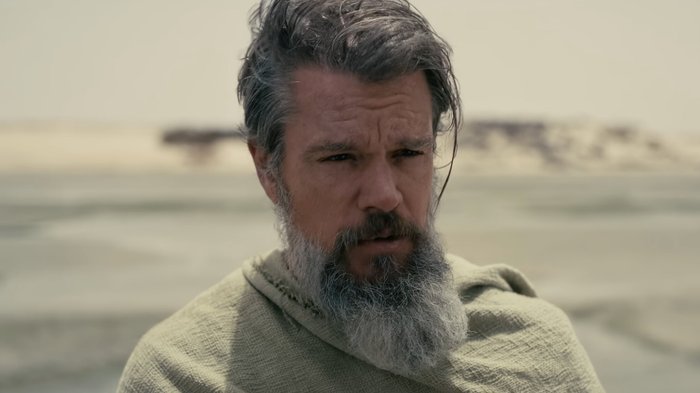 Man with gray beard wrapped in a rough beige cloak stands on a windswept beach, gazing off to the side with a serious expression.