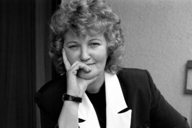 Portrait of a smiling woman with curly hair resting her chin on her hand, wearing a dark blazer and white collar.