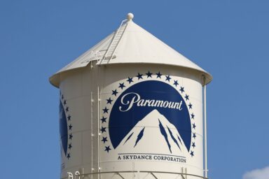 White water tower with the Paramount logo and mountain emblem against a clear blue sky.