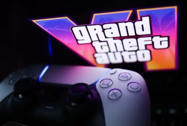 White PlayStation controller in the foreground with a neon Grand Theft Auto sign glowing in the background.