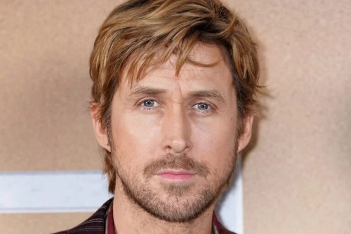 Close-up portrait of a man with blond hair, blue eyes, and light stubble looking at the camera on a beige background.