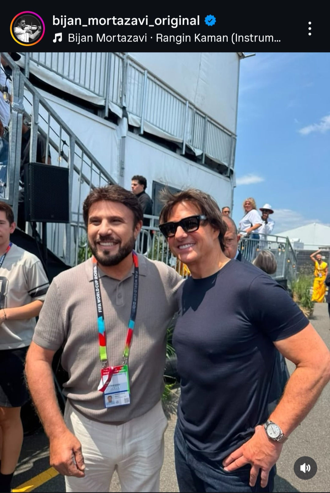 1000105502 Two Men Smile for a Photo Backstage at an Event Wearing Lanyards and Casual Shirts with a Metal Railing and White Building in the Background Under a Blue Sky