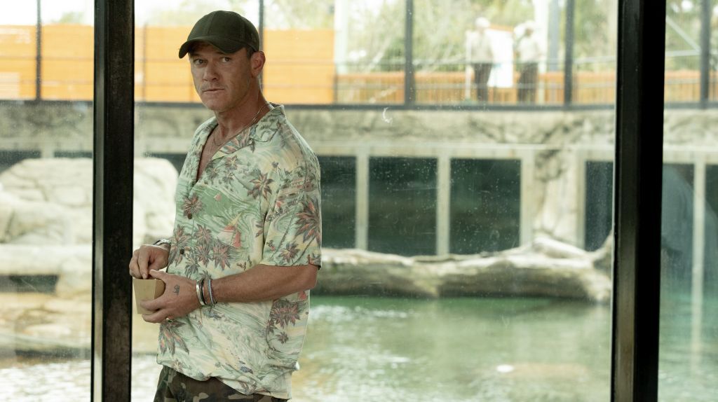 Man in a Tropical Shirt and Cap Standing by a Glass Barrier Holding a Cup