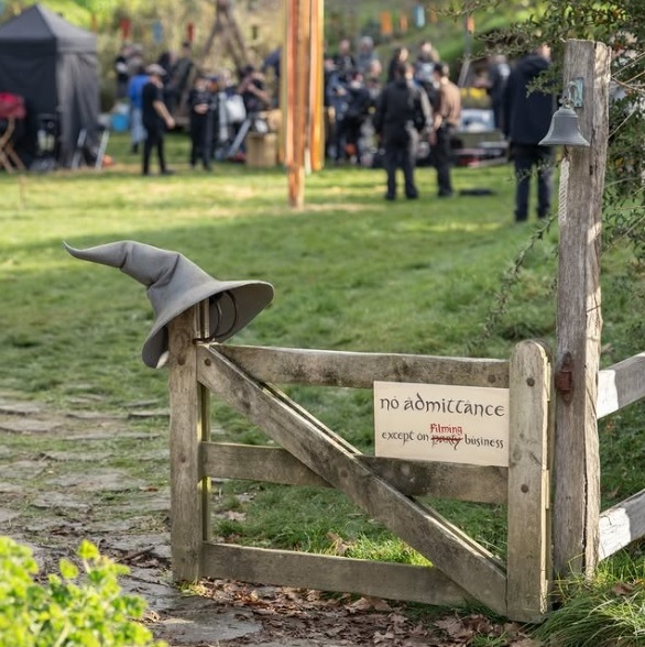 Wooden Gate with a Crooked Witch's Hat on Top a Sign Reads 'no Admittance Except on Filming' As People Gather in the Background at a Film Set