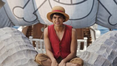 Young man in a straw hat and red vest sits cross‑legged among large sand sculptures at a beach exhibit.