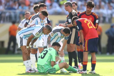 Soccer players in blue-and-white striped jerseys surround a kneeling goalkeeper in a bright green kit on the field during a match moment.