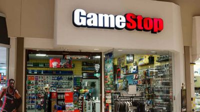 GameStop storefront in a mall, glass doors open to a stocked store with video games and accessories visible inside.