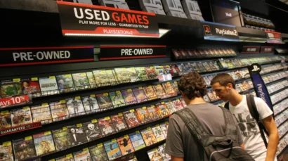 Two customers browse a game store's used/pre-owned section, shelves filled with PS2 titles under bold USED GAMES and PRE-OWNED signs.