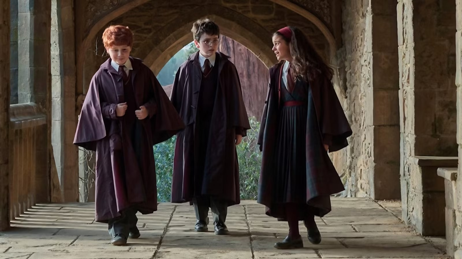 Three Students in Maroon Robes Walk Together Through a Stone Cloister Talking As They Stride Down a Arched Arcade