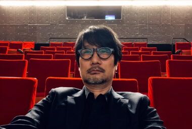 Self-portrait of a man wearing round glasses and a dark suit, seated in an empty red theater auditorium.