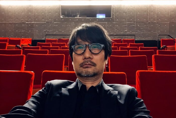 Self-portrait of a man wearing round glasses and a dark suit, seated in an empty red theater auditorium.