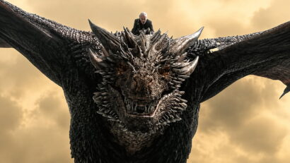 Dark dragon with jagged scales and orange eyes, a man in black perched on its head as it soars against a yellow, cloudy sky.