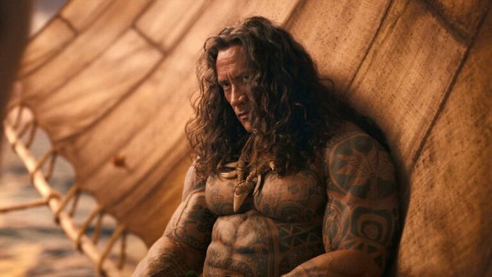 Shirtless, heavily tattooed man with long hair rests against a large brown sail on a sunny beach, wearing a bone necklace.