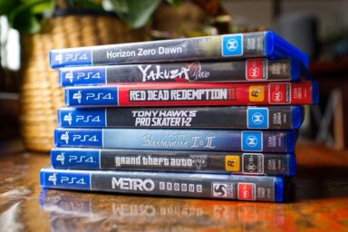 Stack of PS4 game cases on a wooden table, showing Horizon Zero Dawn, Yakuza Kiwami/Zero, Red Dead Redemption II, Tony Hawk's Pro Skater 1+2, Shenmue I & II, Grand Theft Auto V, and Metro Exodus.