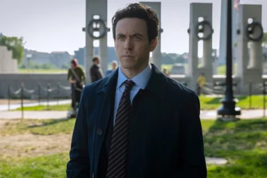 Man in a navy coat and striped tie stands outdoors with modern columns in the background.