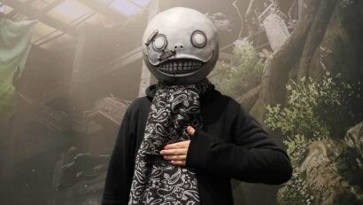 Person in a black hoodie wearing a large gray mask with round eyes and a stitched mouth, with a paisley scarf, hand on chest.