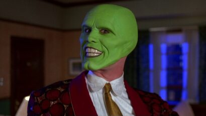 Green-skinned person in a red patterned suit, wearing a white shirt and gold tie, with a wide toothy grin indoors.