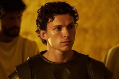 Young man in dark armor gazes to the right in a warm, amber-lit scene.