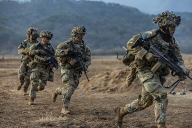 Four soldiers in camouflage fatigues and ghillie helmets sprint across a dry, open field during a training exercise, rifles ready.