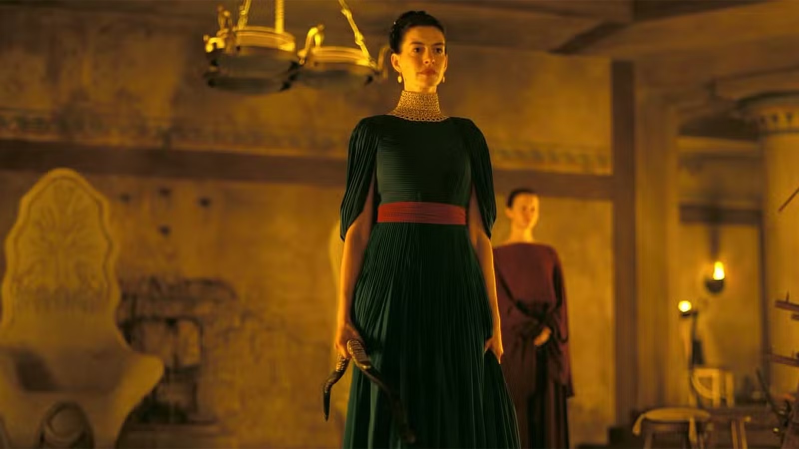 Woman in an Emerald Green Pleated Gown with a Red Sash Stands in a Dim Amber lit Antique Room Holding a Dagger at Her Side As Another Person Watches from Behind