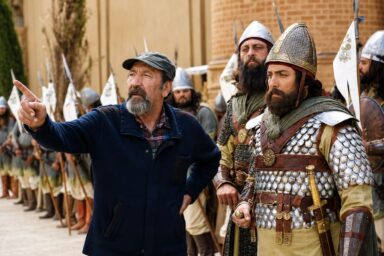 Older man with gray beard points to the left, surrounded by medieval soldiers in armor and helmets on a film set