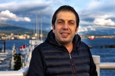 Smiling man in a black quilted vest stands at a harbor railing with water and boats in the background.
