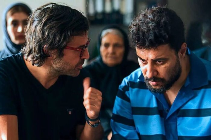 Two men in the foreground have a tense discussion; one with red glasses gestures with a thumbs-up while the other looks down, with a woman in a hijab blurred in the background.