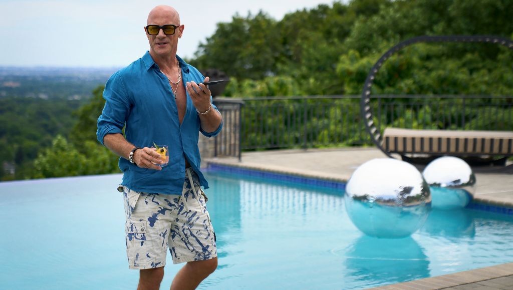 1000020587 bald Man in Sunglasses Blue Shirt and Shorts Stands by a Modern Pool Holding a Drink and Phone with Silver Spheres in Water Lounge Chair and Lush Green Trees in the Background
