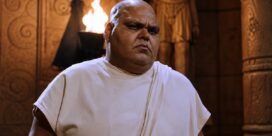 Man with a shaved head in a white robe stands inside a carved stone temple, illuminated by a nearby fire.