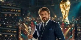 Man in a navy pinstripe suit holds a violin on a bright concert stage, with a large golden trophy in the background and audience lights.