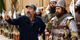 Older man with gray beard points to the left, surrounded by medieval soldiers in armor and helmets on a film set
